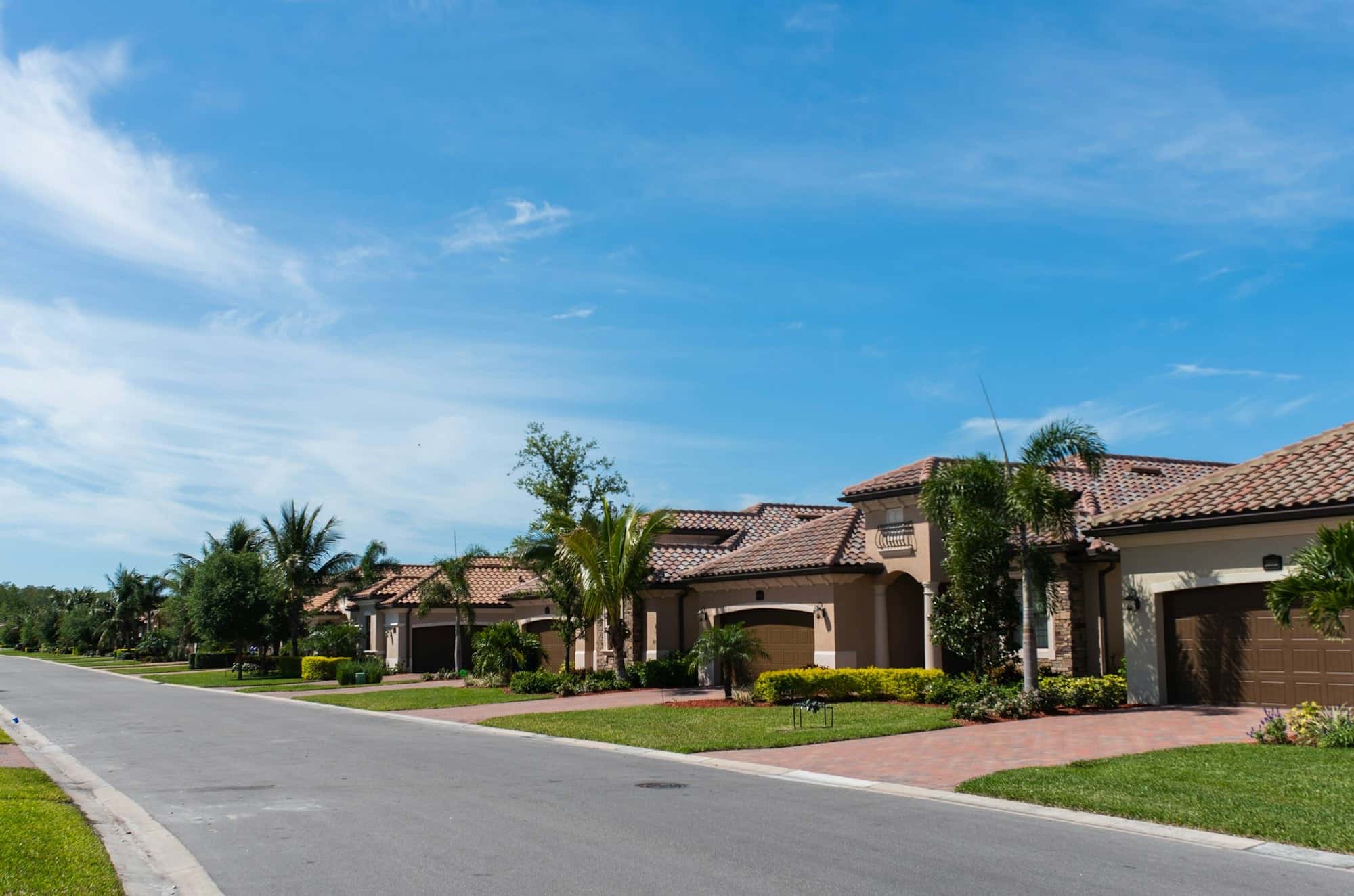Residential street of tile-roofed stucco homes with brick-paver driveways, trimmed shrubs, lawns, and small palms
