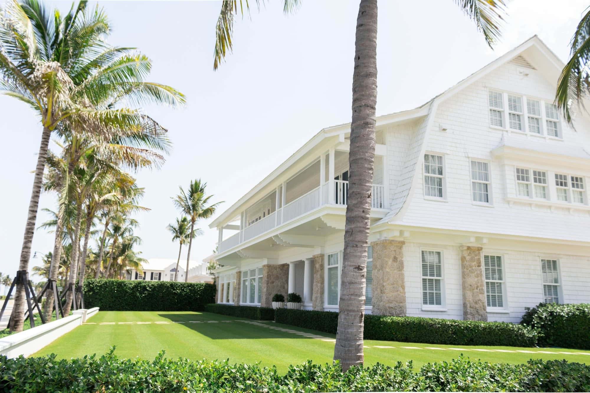 Large white shingle-style house with a striped mown lawn, low boxed hedges along the edges, and coconut palms in front