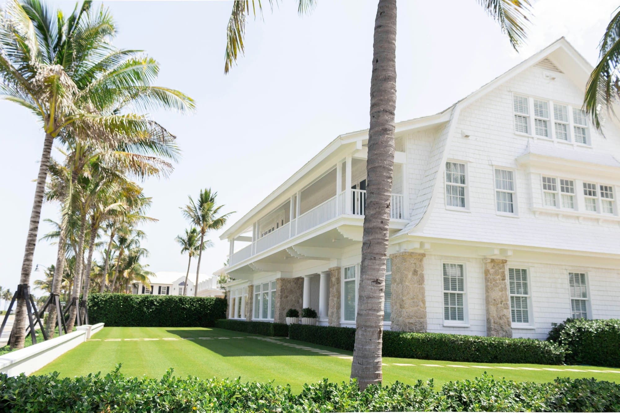 Large white shingle-style house with a striped mown lawn, low boxed hedges along the edges, and coconut palms in front