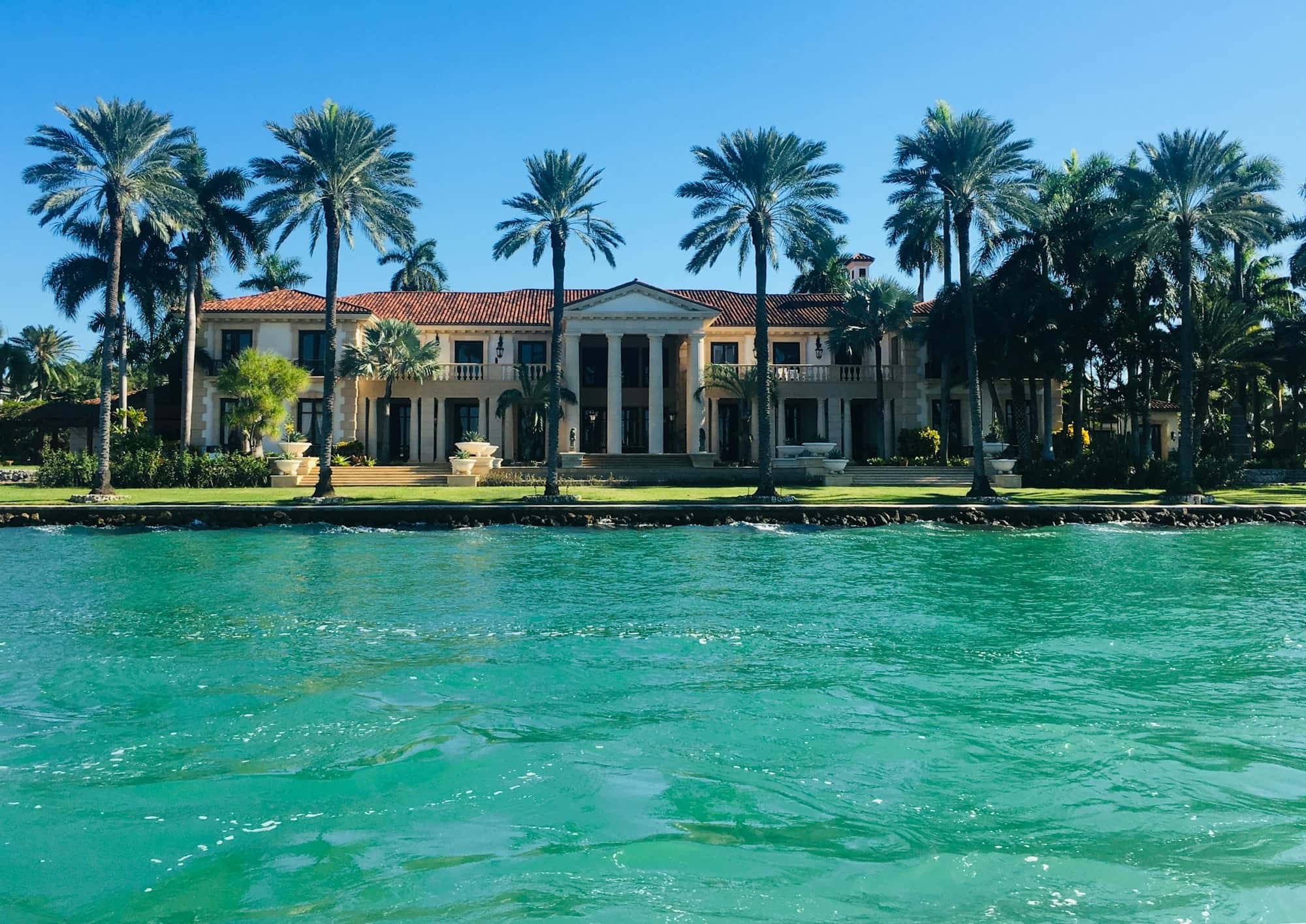 Columned tile-roof mansion behind a seawall and a row of tall palms, seen from turquoise water
